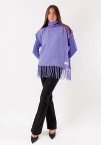 Poncho viola con collo a dolcevita e bordo frangiato, indossato sopra un top a maniche lunghe, abbinato a pantaloni neri svasati e ballerine a punta.