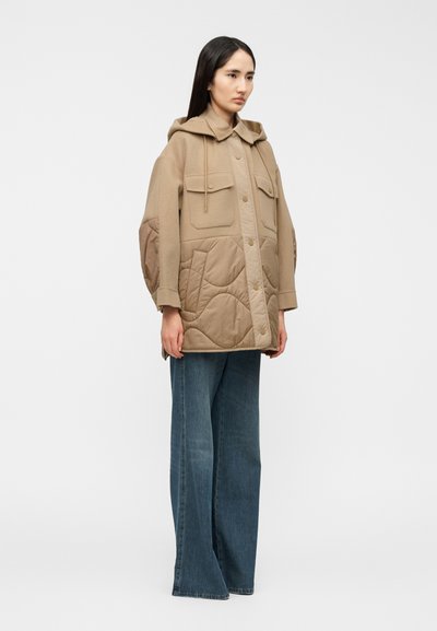 Μπεζ φούτερ με κουμπιά και δύο τσέπες στο στήθος, με επενδύσεις σε στυλ κοντό ραφής. Συνδυάζεται με τζιν παντελόνι φαρδύ από κάτω.