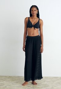 Črn bikini zgornji del s prednjo vozlano podrobnostjo v kombinaciji s črnimi drapiranimi širokimi hlačami. Gladka tekstura, lahka materiala, minimalistična zasnova.