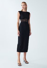 Robe en dentelle noire avec une silhouette cintrée, des manches courtes, une incrustation en dentelle transparente à la taille et un ourlet arrivant au genou. Portée avec des sandales à talons noires.