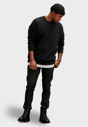 Sort sweatshirt med tekstureret logo mønster, lagd oven på en hvid T-shirt, parret med sorte bukser og sorte snørebånds kampstøvler.