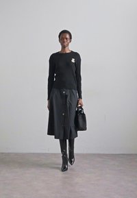 Modelis stāv pārliecināti melnā ribotā džemperī un garā svārkā, turot roku elegantu melnu rokas soma pret vienkāršu fona.