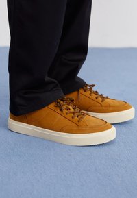 Baskets en daim marron avec semelles en caoutchouc blanches, dotées de lacets et de coutures marron. Portées avec un pantalon noir sur une surface bleue.