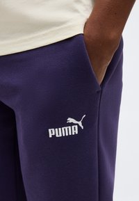 Κοντινό πλάνο ενός ατόμου με το χέρι στην τσέπη, που φοράει σκούρο μπλε παντελόνι Puma και ανοιχτό μπεζ πουκάμισο ορατό στη μέση.