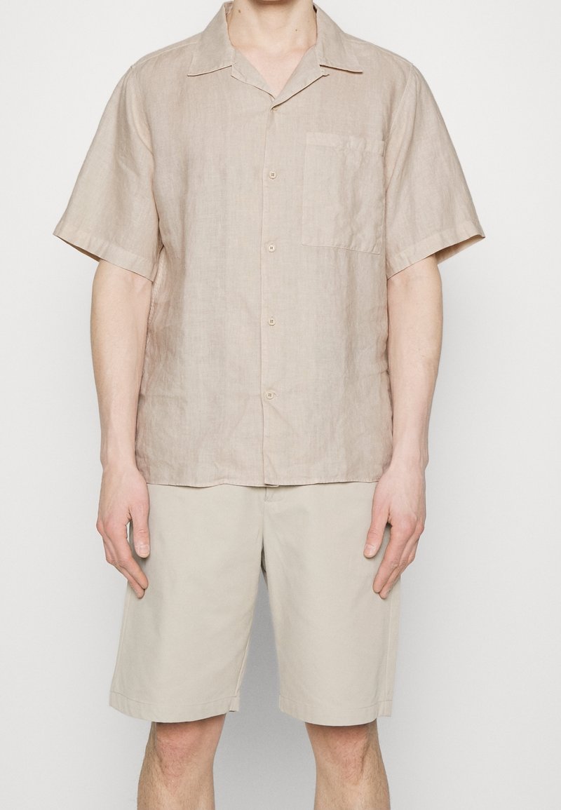 Lichtbruine linnen korte mouw shirt met een zak, kraag en knoopsluiting. Gecombineerd met beige op maat gemaakte shorts, beide casual en relaxed pasvorm.
