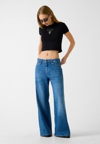 Czarny krótki t-shirt z logo "GUESS" w kolorze złotym, zestawiony z jasnoniebieskimi szerokimi spodniami z zapinaniem na guziki i szlufkami na pasek.