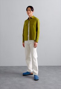 Olivnozeleni zavihek s kratkimi rokavi s dvema žepoma na prsih, pod njim bela majica, bele hlače in modri čevlji s črnimi podplati.