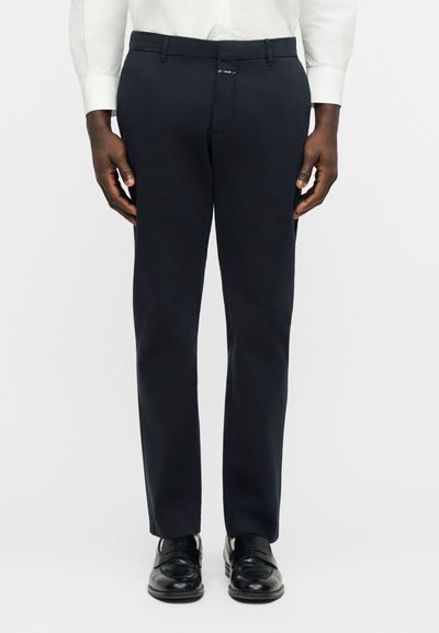 Homme portant un pantalon habillé noir, une chemise blanche et des chaussures en cuir noir, debout devant un fond clair uni.