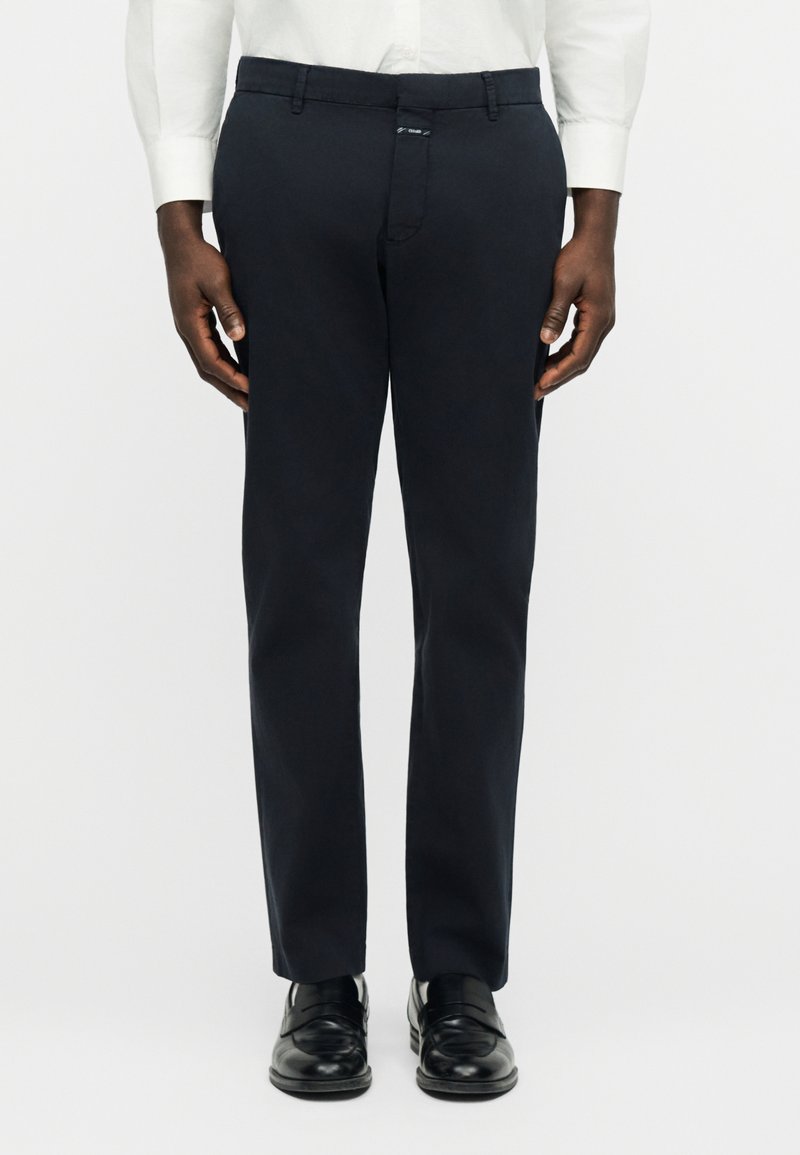 Homme portant un pantalon habillé noir, une chemise blanche et des chaussures en cuir noir, debout devant un fond clair uni.