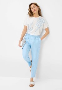 White t-shirt with blue abstract pattern, paired with light blue cargo pants. Model holds a metallic clutch, wearing white sandals.