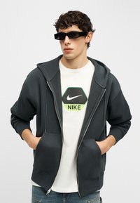 Νεαρός άνδρας που φοράει μαύρα γυαλιά ηλίου, μαύρη ζακέτα με φερμουάρ, λευκό T-shirt με λογότυπο Nike και τζιν, ποζάροντας με τα χέρια στις τσέπες της ζακέτας.