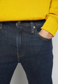 Pantalones vaqueros de mezclilla azul oscuro con costuras visibles y un bolsillo frontal. Llevados con una sudadera amarilla brillante en la cintura.