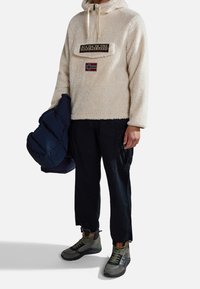 Kremowa bluza z polaru z pół zamkiem, naszywką z logo i norweską flagą; w połączeniu z ciemnymi spodniami cargo i wielokolorowymi wysokimi butami. Trzyma granatową kurtkę puchową.