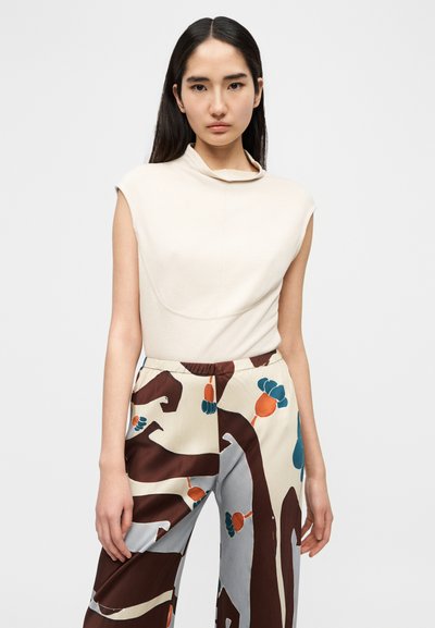 Beige ermeløs topp med oppbrettet krage, kombinert med mønstrede, flerfargede bukser med abstrakte design i brunt, blått og oransje.