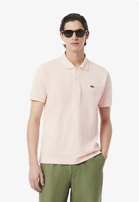 Ljust rosa polo skjorta med klassisk krage, korta ärmar och en grön krokodil-logotyp på vänstra bröstet, kombinerad med gröna byxor.