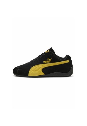 Schwarzer Puma-Sneaker mit gelbem Seitenstreifen, Puma-Logo, schwarzen Schnürsenkeln und strukturierter schwarzer Sohle, seitlich auf weißem Hintergrund gezeigt.