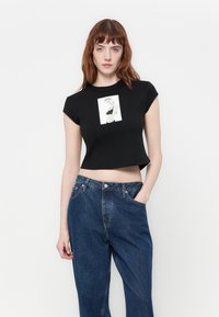 Zwarte cropped t-shirt met kapmouw en een grafische print aan de voorkant. Gecombineerd met loszittende blauwe denim broek. Glad stoffen textuur.