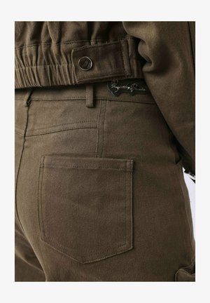 Bruine katoenen broek met een hoge tailleband, achterzakken en een metalen knoopdetail. De stof heeft een gladde, gestructureerde afwerking.