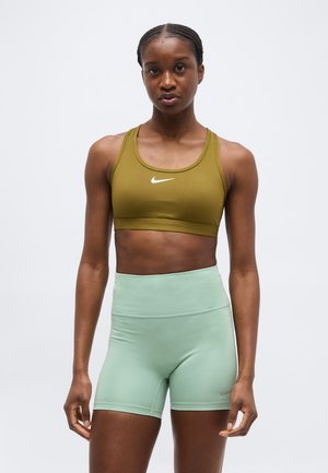 Donna atletica in piedi, indossa un reggiseno sportivo oliva e pantaloncini da ciclismo a vita alta verde chiaro, rivolta in avanti contro uno sfondo semplice.