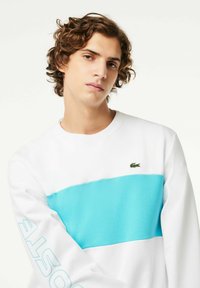 Sudadera blanca con una franja horizontal azul, presenta un pequeño logo verde. Textura suave, cuello redondo y detalles impresos en la manga.