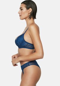 Vrouw met nat haar die een bijpassende blauwe kanten bh en slipje draagt, staande in profiel tegen een effen witte achtergrond.