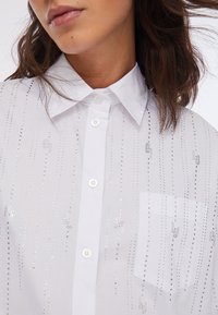 Chemise blanche boutonnée avec des détails en perles argentées et une poche poitrine gauche. Tissu lisse avec un col classique et une fermeture à boutons.