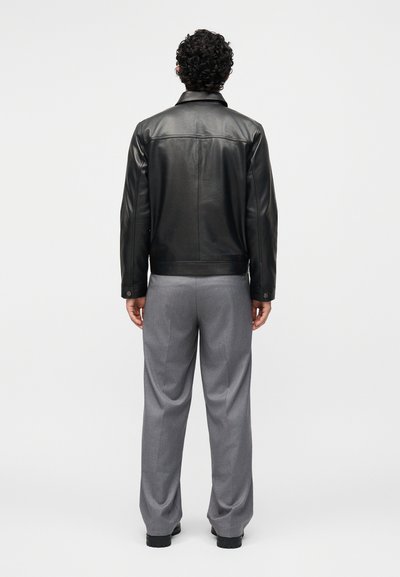 Άνδρας με σγουρά μαλλιά που φοράει μαύρο δερμάτινο μπουφάν και γκρι παντελόνια, στέκεται κοιτάζοντας ένα απλό λευκό φόντο.