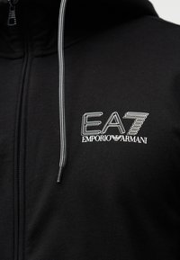 Μαύρο φούτερ με φερμουάρ κατασκευασμένο από απαλό ύφασμα, με εμφανές λευκό λογότυπο EA7 Emporio Armani και αντιθετικές λεπτομέρειες κορδονιού.
