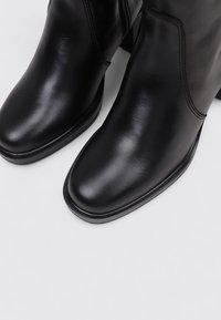 Botines de cuero negro hasta el tobillo con una textura suave, punta cuadrada y cremallera lateral. Presentan un sutil detalle de costura y un tacón bajo y grueso.