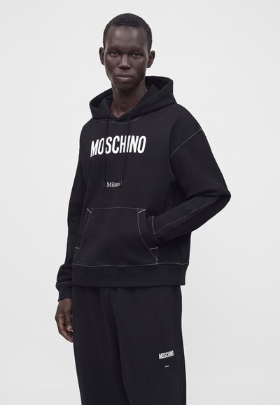 Άνδρας που φοράει μαύρο hoodie Moschino και αντίστοιχο μαύρο παντελόνι με λευκό λογότυπο, στέκεται μπροστά σε ένα απλό ανοιχτό φόντο.