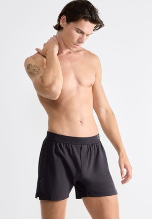 Schwarze Sportshorts mit einem elastischen Bund, die einen leichten Glanz, seitliche Schlitze und dezente Branding auf dem linken Bein aufweisen.