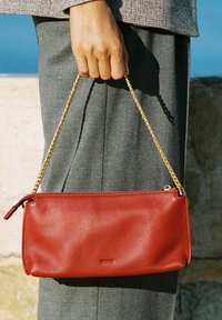 Rdeča usnjena torbica z teksturirano površino, ki ima zlat obročasti trak in zadrgo. Drži jo roka blizu svetlega površja.