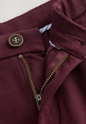 Pantalons bordeaux avec un zip sur le devant et une fermeture éclair en laiton, dotés d'un bouton en relief bien visible et d'une ceinture intérieure rayée contrastante.