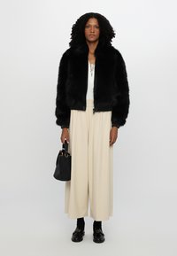 Bomber jacket in pelliccia sintetica nera, polsini a costine, top bianco con lacci, pantaloni beige a gamba larga, borsa nera e scarpe nere chunky.