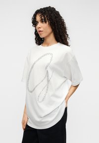 Άσπρο βαμβακερό μπλουζάκι με μεγάλο ασημί λαμπερό σχέδιο 'G'. Χαλαρή εφαρμογή με κοντά μανίκια και στρογγυλή λαιμόκοψη, σε συνδυασμό με σκούρο παντελόνι.