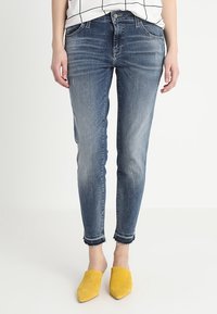 Mujer vestida con unos pantalones vaqueros ajustados de color azul desgastado con bajos deshilachados y zapatos amarillos de punta con diseño deslizante, de pie contra un fondo blanco.