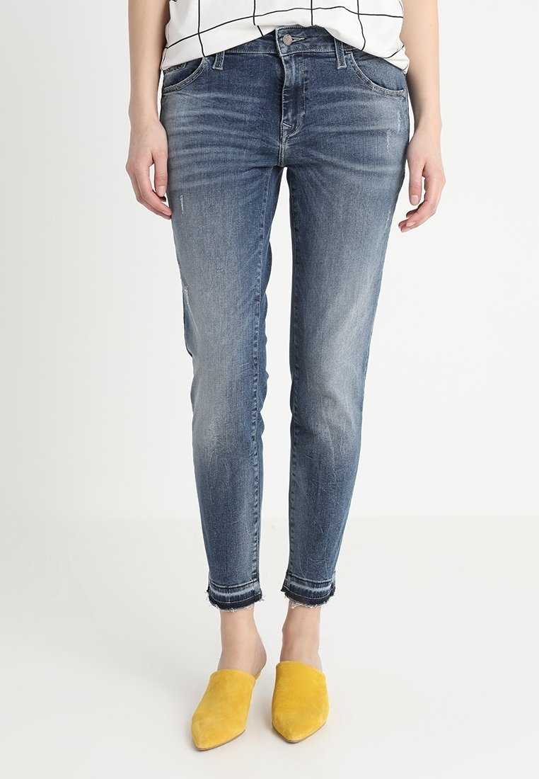 Mujer vestida con unos pantalones vaqueros ajustados de color azul desgastado con bajos deshilachados y zapatos amarillos de punta con diseño deslizante, de pie contra un fondo blanco.