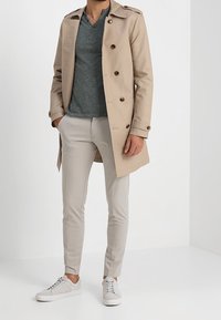 Mężczyzna w beżowym trenczu, szarym swetrze w serek, jasnobeżowych wąskich spodniach i białych trampkach stoi z rękami w kieszeniach.