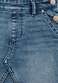 Denim tkanina svetlo modre barve. Vsebuje visoko pasnico, dva bakrena gumba in opazne šivalne vzorce po vsej površini.