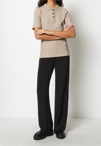 Beige ribbet polo skjorte med korte ærmer og tre sorte knapper, parret med løstsiddende sorte bukser og robuste sorte sko.