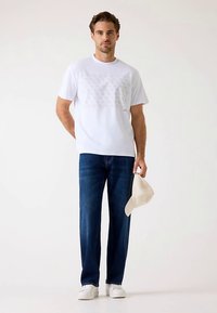 T-shirt bianca con stampa geometrica sottile, abbinata a jeans blu scuro. Modello tiene un cappellino beige, indossa sneakers bianche. Outfit casual.