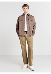 Braune Button-Up-Hemd mit zwei Brusttaschen, weißem T-Shirt darunter, beigen Hosen und weißen Sneakers mit grauen Akzenten.