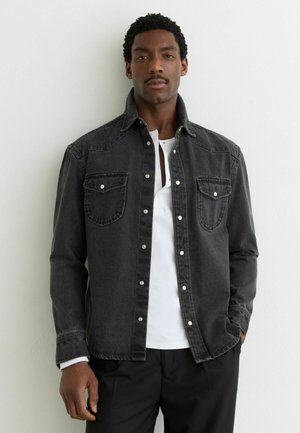 Mand iført en sort denim-knapjakke over en hvid skjorte, stående mod en ensartet lys væg med den ene hånd i lommen.