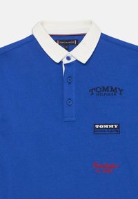 Polo shirt bleu en tissu lisse, avec un col blanc, deux boutons bleus et des logos de marque brodés en bleu marine et rouge.