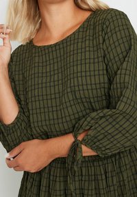 Olivi vihreä mekko, jossa on teksturoitu kangas, ruudukkomalli, 3/4-hihat ja solmittavat koristeet hihoissa.