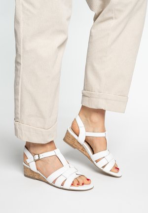 Feet wearing white strappy wedge sandals with cork heels, paired with light beige rolled-up corduroy pants.