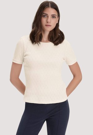 Junge Frau mit schulterlangem braunem Haar, die ein weißes, strukturiertes Kurzarmoberteil und dunkle Hosen trägt, vor einem schlichten Hintergrund stehend.