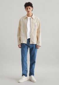 Beige katoenen jack met een kraaghalslijn, borstzak, twee onderste zakken; gecombineerd met blauwe spijkerbroeken en witte sneakers.