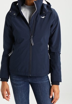 Persoon draagt een marineblauwe jas met rits en capuchon over een grijs shirt en een blauwe spijkerbroek, handen ontspannen langs het lichaam.
