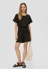 Blouse en lin noir à manches larges et short assorti, noué à la taille. Le modèle tient un sac en osiers crème et porte des sandales à plateforme noires.